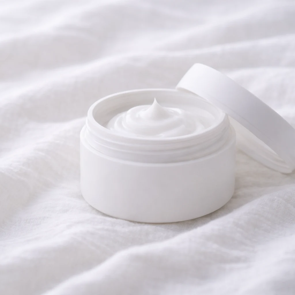 白い布の上に、白いクリームジャーが静かに置かれた清潔感のあるケアのイメージ。
